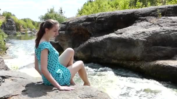 Belle fille a un repos assis sur une pierre près d'une rivière de montagne. Elle mouille ses pieds et joue avec l'eau . — Video