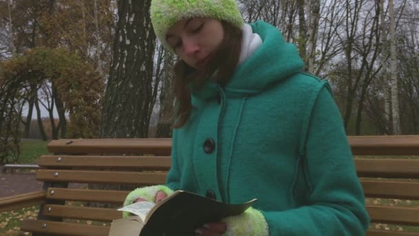 Una joven lee la Biblia en el parque sentada en el banco. El frío otoño . — Vídeo de stock