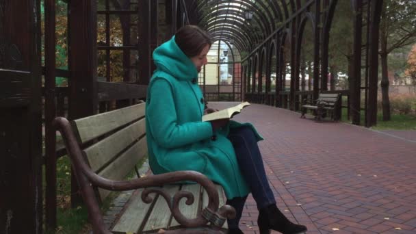 一个年轻的女孩读圣经 》 在公园里坐在长椅上。寒冷的秋天. — 图库视频影像