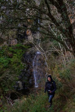 Arka planda şelalesi olan ağaçların arasında kış şapkası takan genç ve güzel beyaz beyaz bir kadının portresi.