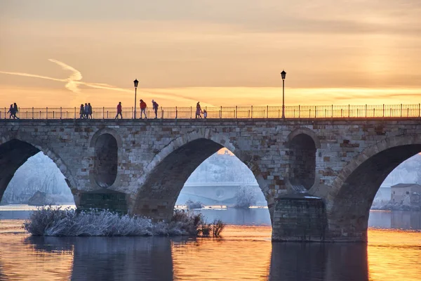 People walking on a stone bridge at sunset and the frozen trees in winter at the golden hour. Stone arches, reflection of the sun on the river. Two bridges. Sewers in the background. Horizontal view