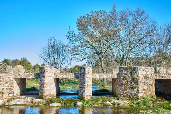 Kırsal kesimde küçük bir dere boyunca uzanan taş köprü. Arkasında açık bir bahar gökyüzü ve ağaçlar var..
