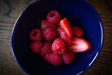 Ahşap masaya konmuş ahududu ve çilekle dolu koyu mavi bir kase..
