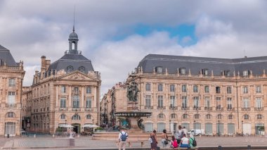 Place de la Bourse zaman atlaması Fransa 'nın Bordeaux şehrinde, Bulutlu mavi gökyüzünün altında klasik mimari ile çevrili Üç Zarafet Çeşmesi ile birlikte. İnsanlar ana meydanda bir tramvay durağında