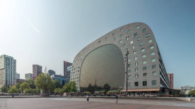 Rotterdamse Markthal 'ın dış yüzeyinde aşırı kayma. Dükkanları ve restoranları olan ikonik at nalı şeklinde bir bina. Meydanda gezinen insanlar. Gökdelenler ve arka planda canlı bir şehir manzarası