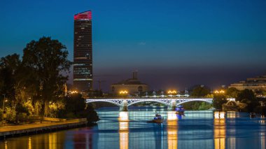 Guadalquivir Nehri yakınlarındaki Torre Sevilla gökdeleninin yer aldığı Seville 'in ufuk çizgisi gece gündüz geçiş zamanı. Aydınlatılmış Triana Köprüsü su, modern ve geleneksel mimariyi yansıtıyor