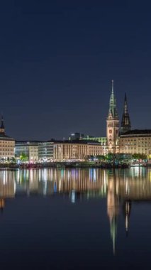Binnenalster 'in Hamburg, Almanya' daki Belediye Binası ve Nikolai Kilisesi 'ndeki gece programı. Aydınlatılmış tarihi binalar yıldızlı bir gökyüzünün altındaki Alster Gölü sularına yansıyor. Manzaralı şehir manzarası