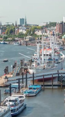Almanya 'nın Hamburg kentinde Elbe nehir kenarının hava zaman çizelgesi. İskeleye yakın tekneler, Jan Fedder gezinti güvertesi, marina ve konteyner gemilerinin geçtiği manzara. Elbe Nehri kıyısındaki canlı şehir manzarası