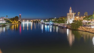 İspanya, Seville 'deki Torre del Oro gözlem kulesi yakınlarındaki Guadalquivir Nehri boyunca yüzen bir tur teknesi. Günbegün geçiş zamanı, şehrin ışıkları su ve deniz manzarasını yansıtıyor.
