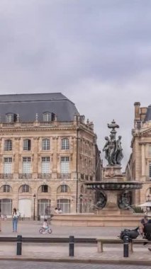 Place de la Bourse zaman atlaması Fransa 'nın Bordeaux şehrinde, Bulutlu mavi gökyüzünün altında klasik mimari ile çevrili Üç Zarafet Çeşmesi ile birlikte. İnsanlar ana meydanda bir tramvay durağında