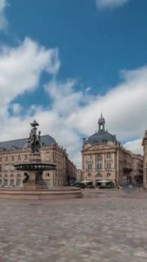 Place de la Bourse zaman atlaması Fransa 'nın Bordeaux şehrinde, Bulutlu mavi gökyüzünün altında klasik mimari ile çevrili Üç Zarafet Çeşmesi ile birlikte. Meydanda yürüyen insanlar.