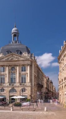 Place de la Bourse, Fransa 'nın Bordeaux şehrinde, bulutlu mavi gökyüzü altında klasik mimari ile çevrili restoranları olan bir yer. Meydanda ve ana caddelerde yürüyen insanlar