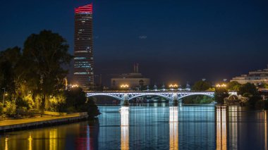 Guadalquivir Nehri yakınlarındaki Torre Sevilla gökdeleninin yer aldığı Seville 'in ufuk çizgisi gece gündüz geçiş zamanı. Aydınlatılmış Triana Köprüsü su, modern ve geleneksel mimariyi yansıtıyor