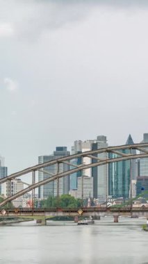 Frankfurt 'un hava zaman çizelgesi Deutschherrnbrucke Demiryolu Köprüsü' nün kemerinden geçiyor. Ana Nehir 'de yüzen botlar ve su yansımaları var. Bulutlu gökyüzü sahneyi çerçeveliyor. Almanya