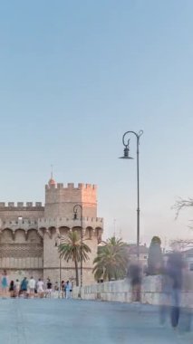 Serrans Kuleleri Torres de Serranos zamanapse Valencia, İspanya 'da. Şehrin antik duvarlarından gelen büyük bir ortaçağ kapısı. İnsanlar gün batımından önce tarihi bir dönüm noktasında akşam gökyüzünün altında bir köprüde yürüyorlar.