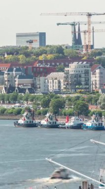 Almanya 'nın Hamburg kentinde Elbe nehir kenarının hava zaman çizelgesi. İskeleye yakın tekneler, marina ve geçen gemilerle manzara. İnşaat alanında vinçleri olan evler. Elbe Nehri kıyısındaki canlı şehir manzarası