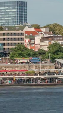 Hamburg limanına giriş ve çıkış yapan bir feribotun rıhtımdaki yolcuları ve merdivenleri gösteren zaman çizelgesi. Arka planda Elbe nehri boyunca tarihi ve modern binalar var. Almanya