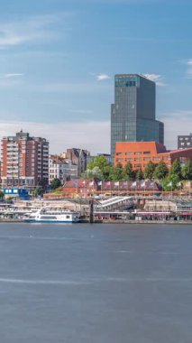 Hamburg limanına giriş ve çıkış yapan bir feribotun zaman çizelgesi. İskelede yolcuları var ve merdivenlerde ilerliyor. Arkaplanda Elbe nehri boyunca tarihi ve modern binalar var. Almanya