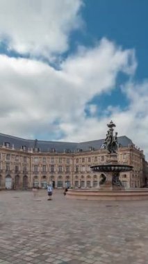 Place de la Bourse zaman atlaması Fransa 'nın Bordeaux şehrinde, Bulutlu mavi gökyüzünün altında klasik mimari ile çevrili Üç Zarafet Çeşmesi ile birlikte. Meydanda yürüyen insanlar.