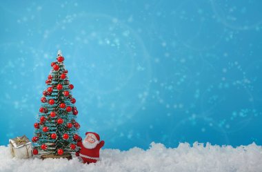 Kırmızı topları olan küçük dekoratif noel ağacı ve beyaz kar üzerinde küçük Noel Baba ile mavi arka plan arasında mesaj için boş yer var..