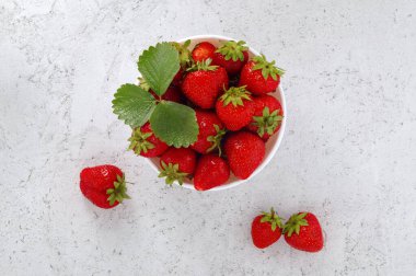 Olgun çilekler beyaz kasede yapraklarla aydınlık beton yüzey, üst manzara.