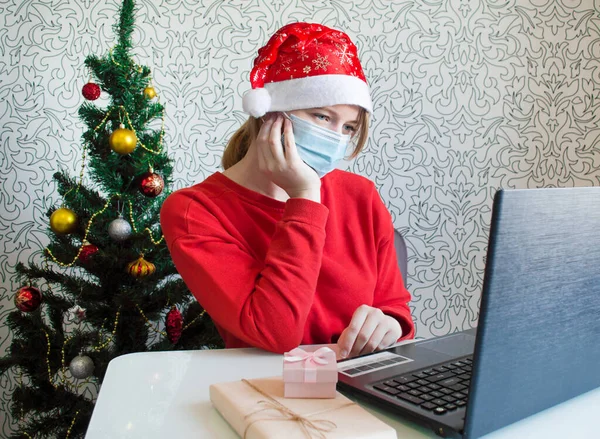 Tıbbi maskeli ve noel hediyeli üzgün genç bir kadın dizüstü bilgisayarına bakıyor. Noel atmosferi. Çevrimiçi iletişim