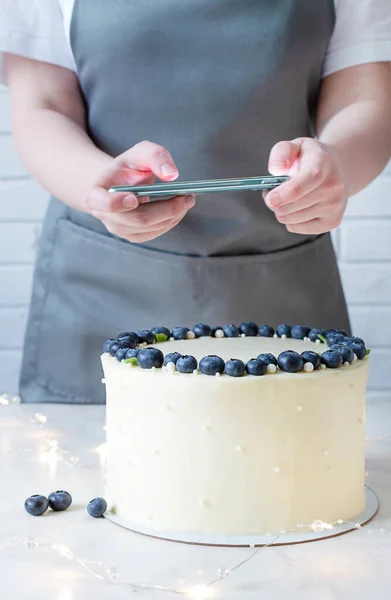 Professional food blogger taking pictures on smartphone. White cake with cream cheese and fresh blueberries