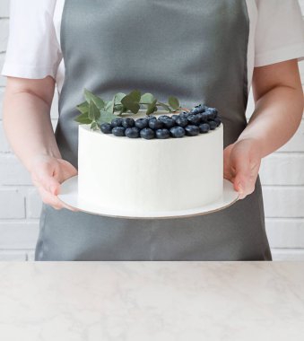 Önlük ve beyaz tişörtlü pasta şefi elinde beyaz bir pasta tutuyor.