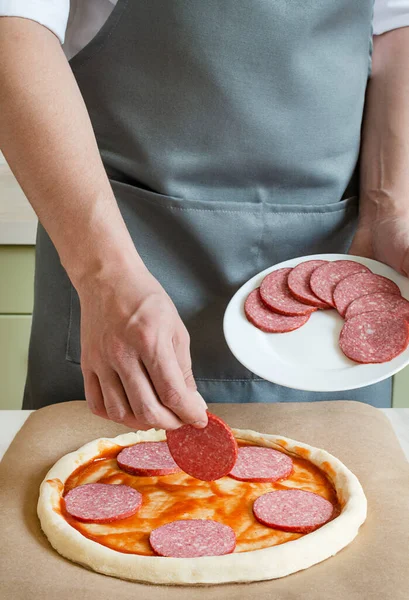 ピザメーカーは 生地に材料を置きます ソーセージとサラミでピザを作る — ストック写真