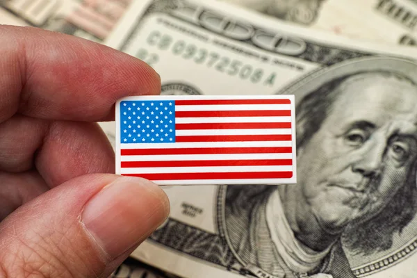 Persons hand holds American Flag badge against heap of dollar bi — Stock Photo, Image