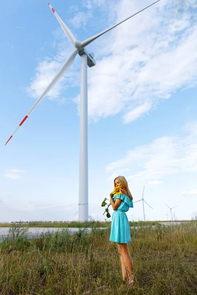 Girl with sunflower in hands against field and wind generators on blurred background