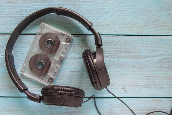 Black headphones and a transparent audio tape lie on a blue wooden background. Space for the text. Top view. Close-up