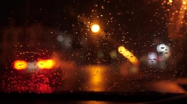 Night street view from the car. It's raining, the streetlights are shining and the traffic lights are blinking. A car passes by. Focus on the windshield