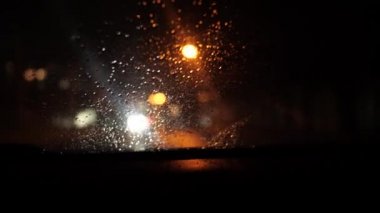 Hareket halindeki bir arabanın ön camından gece şehrinin görüntüsü. Yağmur yağıyor ve sokak lambaları parlıyor. Odaklanma kesiliyor
