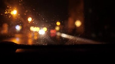 Gece şehri hareket halindeki bir arabanın ön camından. Yağmur yağıyor, ışıklar parlıyor ve yaklaşan arabalar geçiyor. Odaklanma kesiliyor