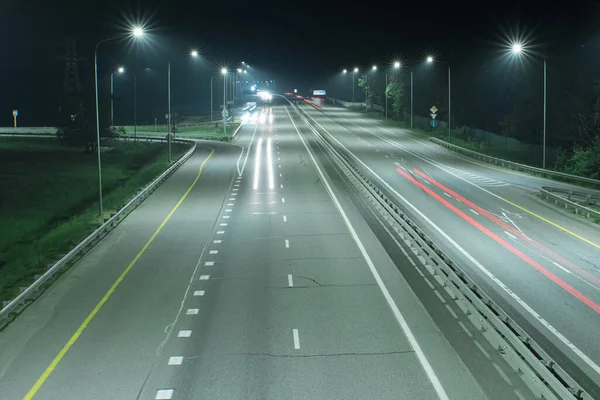 A country highway at night, lit by streetlights and long streaks of light from the headlights of passing cars. Night scenes