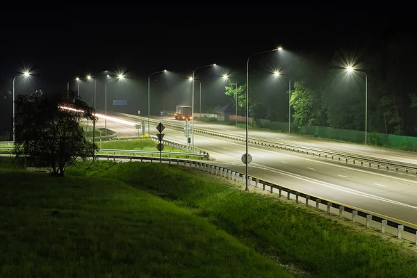 Sokak lambalarının ışığında gece ekspresi. Yolun kenarına park edilmiş bir kamyon var. Yolun kenarında ağaçlar ve yeşil çimenler var. Gece sahneleri
