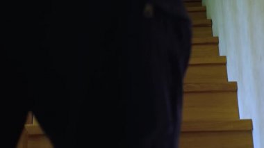 Şortlu ve çoraplı bir adam ahşap bir iç merdivenle ikinci kata çıkıyor. Sabit çerçeve