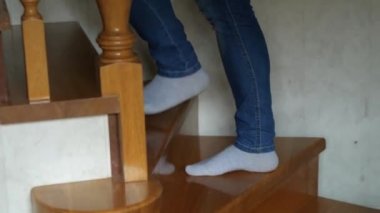 Kot pantolonlu ve beyaz çoraplı ayaklar ahşap bir merdivenin basamaklarında yürür. Yan taraftaki kamera ayakları takip ediyor.