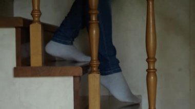 Kot pantolonlu ve beyaz çoraplı ayaklar ahşap bir merdivenden aşağı iniyor. Yan taraftaki kamera ayakları takip ediyor.