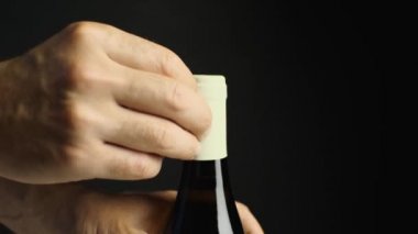 Erkek eli, şarap şişesinin boynundaki ambalajı çıkarır. Yakın plan. Siyah arkaplan