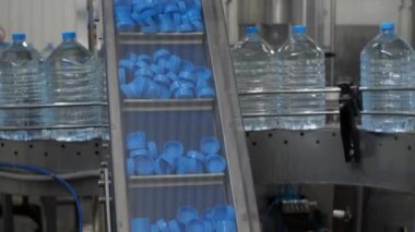 Taşıyıcı bandın üzerindeki mavi plastik kapaklar kaldırılır. Arka planda, 5 litrelik su şişeleri üretim hattı boyunca hareket ediyor. Fabrikada maden suyu üretimi