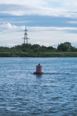 Nehirde kırmızı seyir şamandırası var. Çimenlerin, ormanın ve elektrik kulelerinin arka planında. Mavi günbatımı gökyüzü