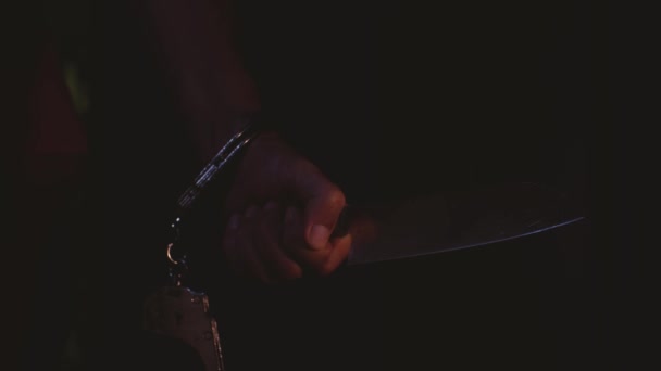 A mans hand cuffed at the wrist holds a large kitchen knife. The blade of the knife glistens in the light and strikes — Stock Video