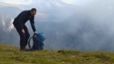 Uçurumun kenarında duran, ekose yün ceketli bir erkek yürüyüşçü sırt çantasını sırtına koyar ve çerçeveden uzaklaşır. Arka planda dağ yamaçları var. Yürüyüş konsepti