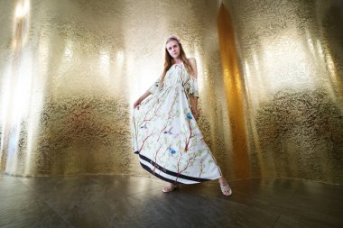 Beyaz elbiseli güzel bir kız uzun bir eteği prenses gibi tutuyor. Tatil için yaz kıyafetleri. Güzel bir iç mekan. Büyük altın duvar. Lüks, refah, zenginlik, petrol ve altın sembolü