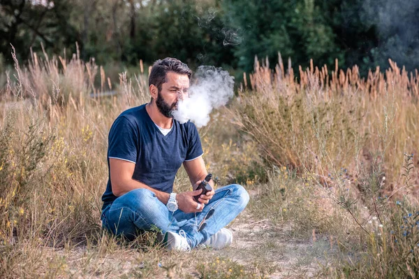 Elegante uomo vaporizzatore barbuto godendo di una sigaretta elettronica nella foresta. La nuova tecnologia sigaretta. — Foto Stock