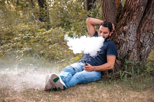 Tarz sahibi sakallı tiryaki orman zemininde birkaç tane e-sigara yaktı. Elektronik sigara.. — Stok fotoğraf