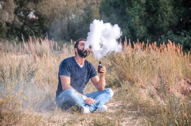 Yakışıklı tiryaki ormanın çimlerinde dinleniyor, buhar çıkarıyor ve bir buhardan buhar çıkarıyor. Vaping konsepti.