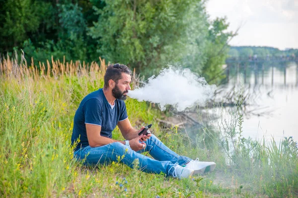 Elegante vaper barbuto fumare una sigaretta elettronica sul terreno della foresta. Modi moderni di smettere di fumare. — Foto Stock